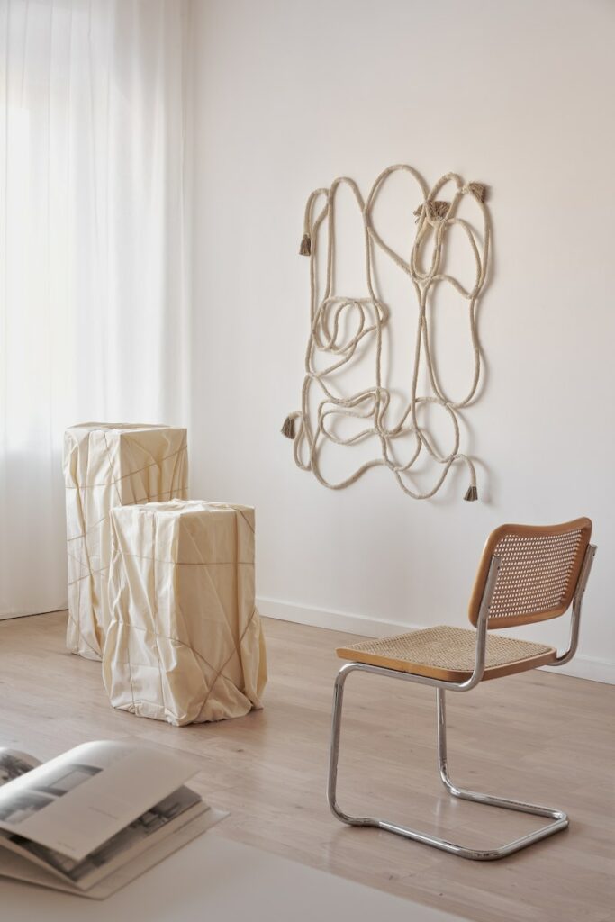 Interior of room with rope decor hanging on wall in room with chair placed near pedestal and opened magazine on table